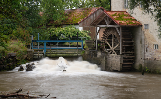 An old historic watermill on the banks of a river. The small waterfall blurs in the long exposure. Trees and plants in the background.