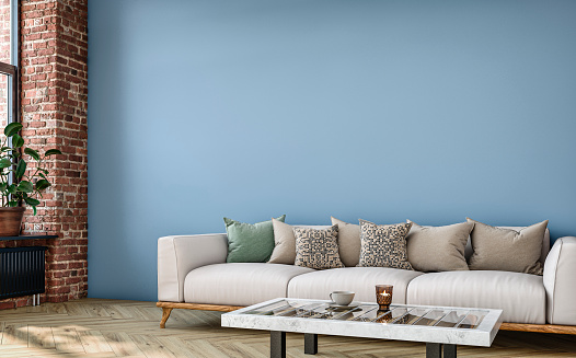 Empty nostalgic retro living room with beige sofa, multi-colored cushions, marble coffee table and decoration (candle, coffee cup) in front of a light blue colored wall background with copy space. A ruined brick wall with a radiator heater under the window on a side and decoration (potted plant ficus ) on the hardwood floor. 3D rendered image.