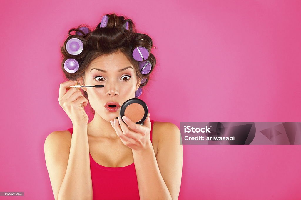 Makeup mascara woman with hair rollers Makeup mascara woman with hair rollers getting ready looking in pocket mirror. Funny image of beautiful funky trendy young mixed race asian caucasian female fashion model putting makeup on pink background. See more: Hair Curlers Stock Photo