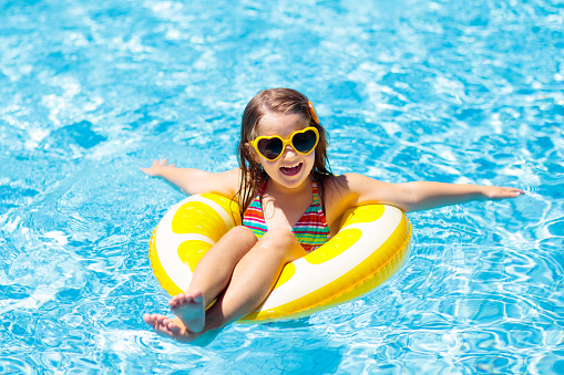Child in swimming pool on inflatable yellow lemon ring. Little girl learning to swim with orange float. Water toy for baby and toddler. Healthy outdoor sport activity for children. Kids beach fun.