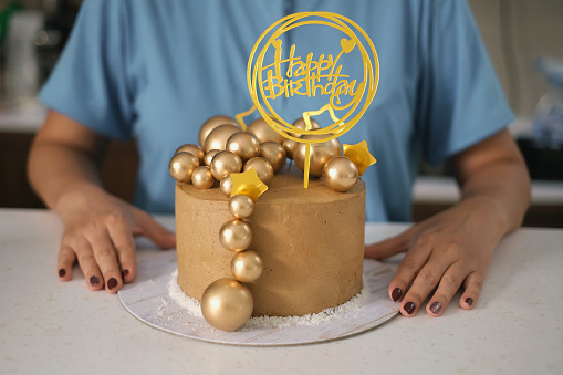 Front view shot of unrecognizable woman sitting in front a birthday cake with gold decoration cake topper