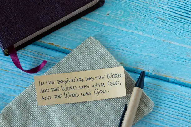 Handwritten biblical text on a note with pen and closed Holy Bible Book on blue wooden background. Top table view.