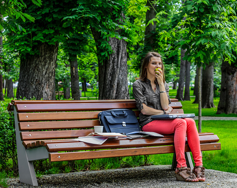 Young woman sitting on a bench in a park and working on a laptop while eating a green apple.