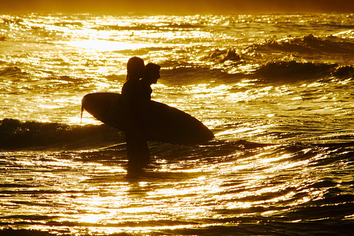 Horizontal seascape photo of two male surfers, in silhouette, carrying surfboards, wading through the water to catch a wave as the sun reflects gold on the rippling water surface. Wategos Beach, Byron Bay, NSW. 23rd November, 2016