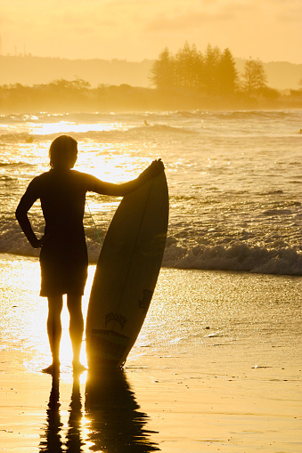 Vertical closeup photo of a young male surfer, in silhouette, holding a surfboard, standing on the beach, looking out over the breaking waves  in the Bay as the sun reflects on the water and wet sand.