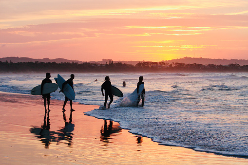 Horizontal landscape photo of four male surfers, carrying surfboards, walking along the beach at sunset as the sunset colors and their silhouettes reflect onto the wet sand.\nThe Pass, Byron Bay, NSW. 23rd November, 2016.