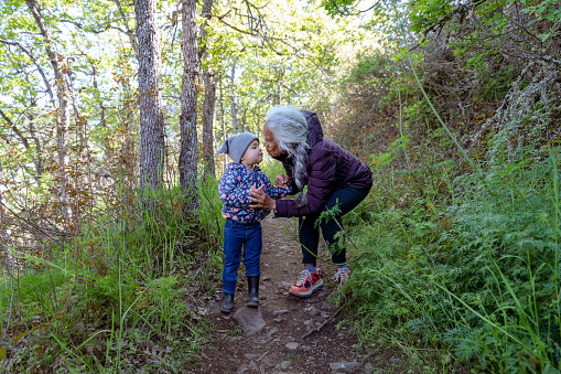 A mixed race senior woman is hiking on a forested trail with her two year old granddaughter. The multi-generation family is staying active and healthy outdoors in Oregon. They are taking a break to give each other a kiss.