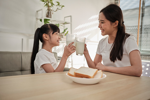 A Healthy Asian Thai family, little daughter, and young mother drink fresh white milk in glass and bread joy together at a dining table in morning, wellness nutrition home breakfast meal lifestyle.