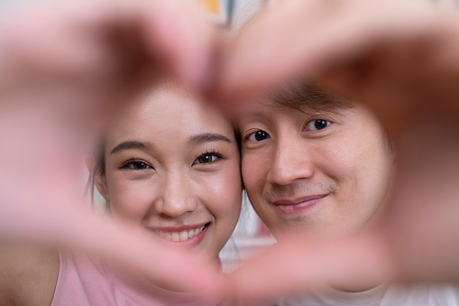 Young Asian couple in a romantic mood making heart-shaped hand gesture. Young couple showing love and affection while playing with camera in home setting.