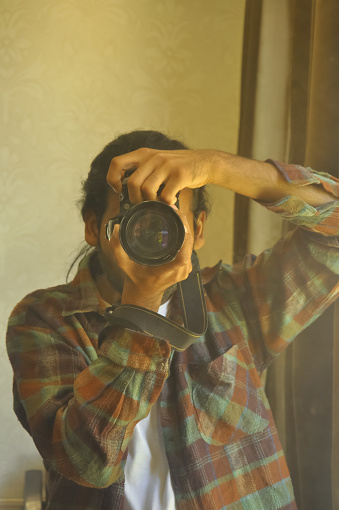 Waist-up of a male photographer looking through the viewfinder of his DSLR camera taking photo of itself front of mirror in the room.