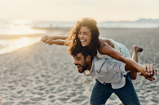 Young playful couple having fun while piggybacking with their arms outstretched on the beach at sunset. Copy space.