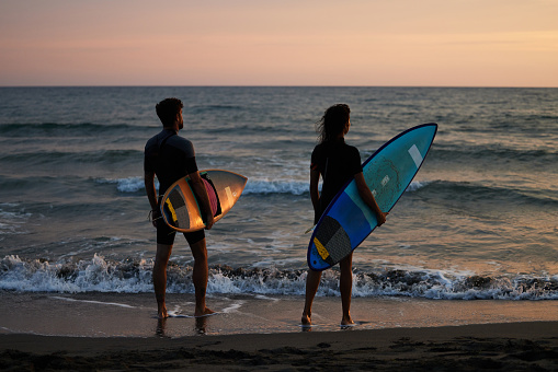 Rear view of couple of surfers standing on the beach with their surfboards and looking at waves at sunset. Copy space.