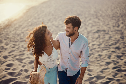 Lovely smiling beautiful woman holding hand of her boyfriend, looking at him, walking on the beach during sunset. Summer island vibes. Love is in the air. Real people lifestyle.