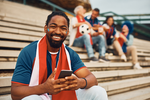 Happy African American sports fan texting on mobile phone outdoors and looking at camera.