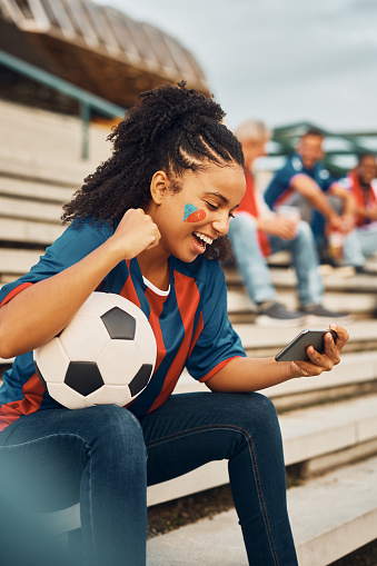 Happy African American woman celebrating while watching football match on mobile phone outdoors.