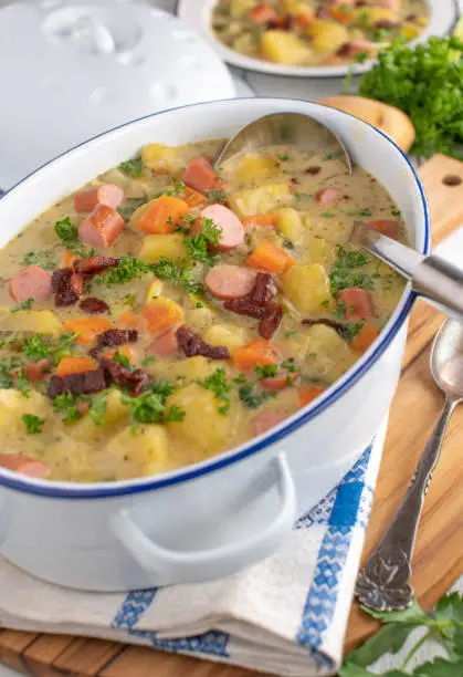Homemade fresh cooked creamy potato soup or potato stew with vegetables, crispy bacon and vienna sausage. Served in a rustic pot with ladle on kitchen table