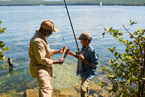 Side view of happy mature black man in activewear showing fish to his grandson with rod while both standing against waterside