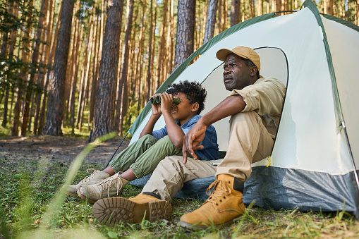 Cute African American boy looking through binoculars while sitting in tent next to his grandfather in casualwear during hike in the forest