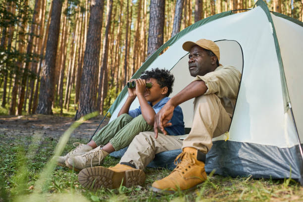 Cute African American boy looking through binoculars while sitting in tent Cute African American boy looking through binoculars while sitting in tent next to his grandfather in casualwear during hike in the forest family camping stock pictures, royalty-free photos & images