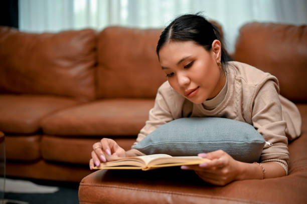 istock Happy and relaxed young Asian female reading a book while lying on her comfortable sofa 1423868096