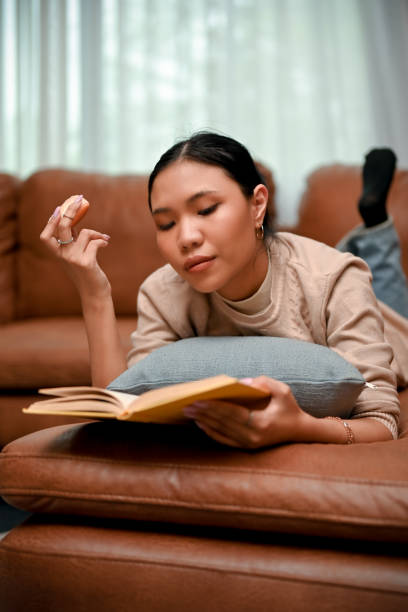istock Portrait, Beautiful Asian female snacking and reading a book while relaxing lying on sofa 1423868108