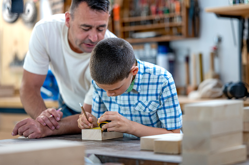 Father observing his son measuring and marking wooden block in workshop.