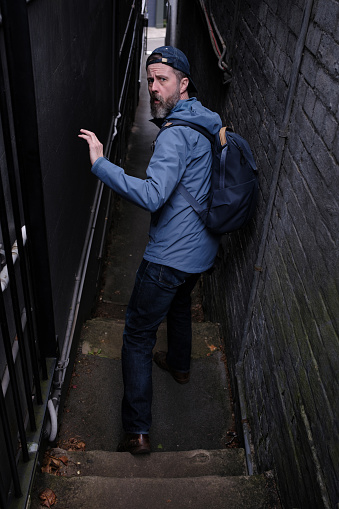 A scared bearded man with cap and backpack looks over his shoulder while running down the stairs of an old dark laneway.