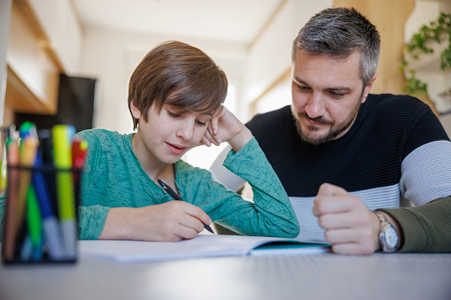 Preadolescent boy sitting at home and doing homework with his father taking care of him, spending time together
