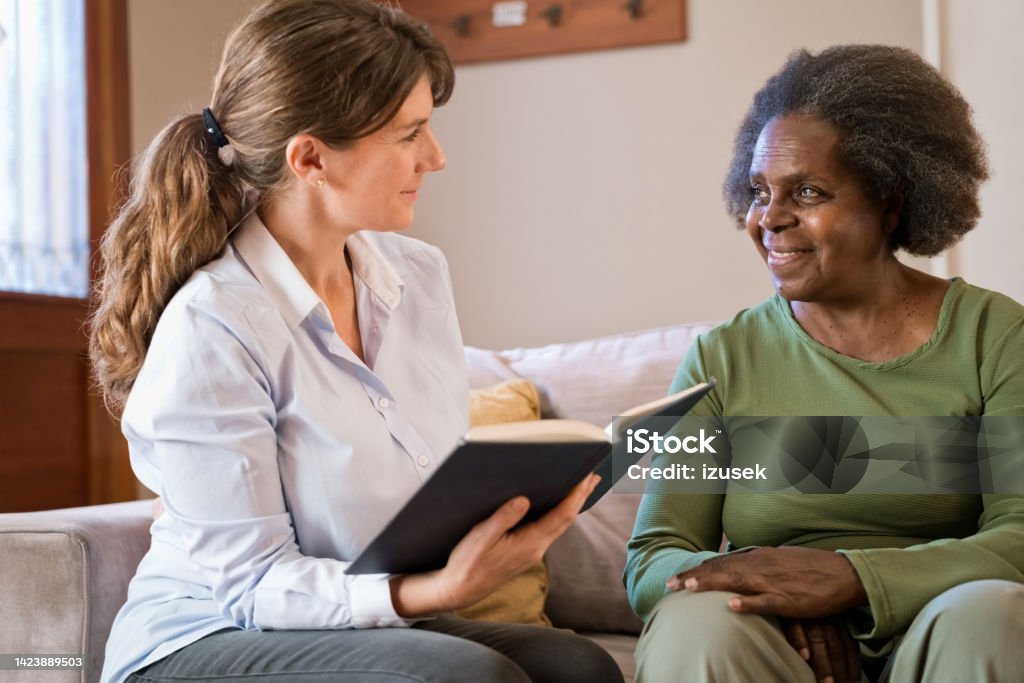 Female nurse reading book for senior woman at home Caregiver reading book for senior woman. Elderly female is sitting by nurse on sofa. They are in living room. Community Outreach Stock Photo