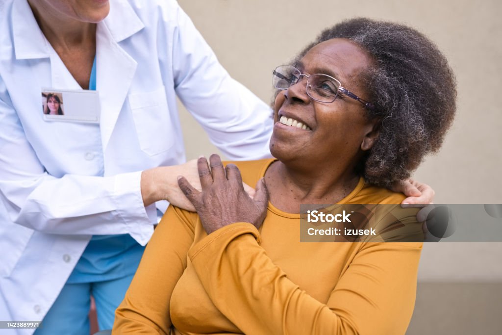 Healthcare worker talking with senior woman Happy female nurse talking with senior woman. Elderly female is looking at caregiver. She is standing against wall. 70-79 Years Stock Photo