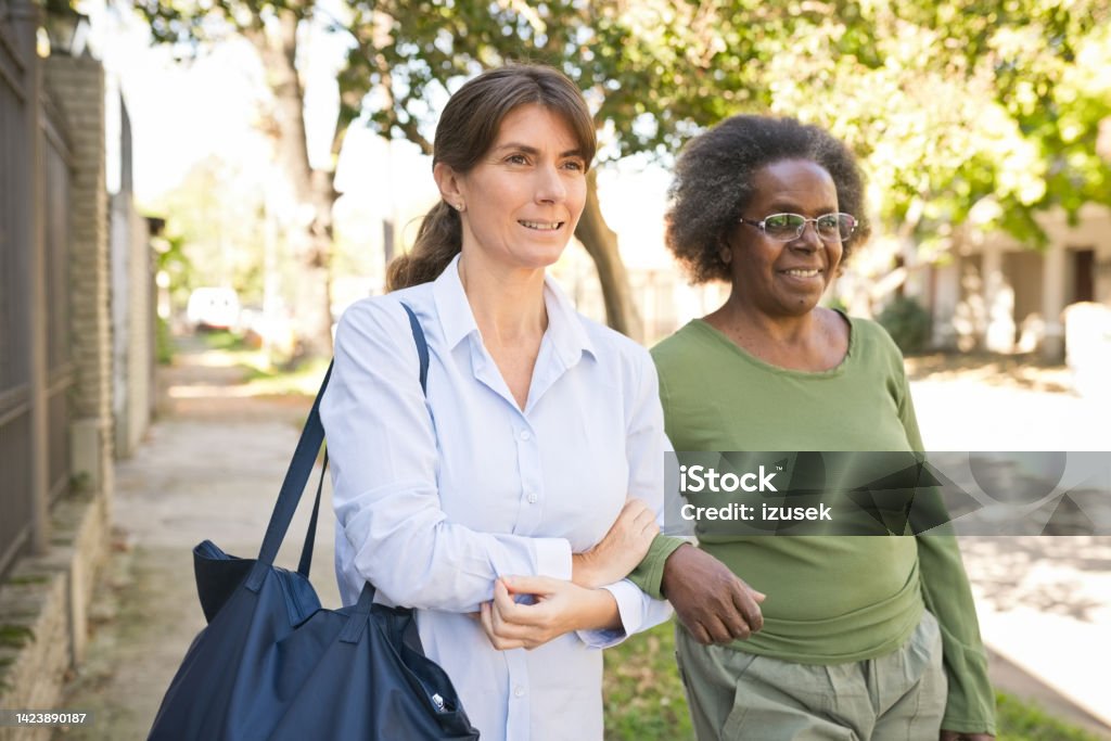 Nurse and senior woman walking on footpath Portrait of caregiver and senior woman on footpath. Nurse and elderly female are walking with arm in arm. Nurse Stock Photo