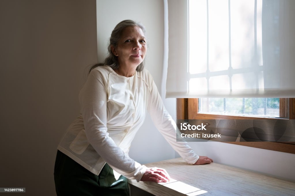 Lonely senior woman in her room Worried elderly woman standing in her room next to the window, looking at camera. 70-79 Years Stock Photo