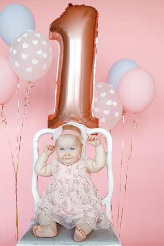 Vertical little joyful, happy, fortunate barefoot one year infant kid in pink dress and flower on head sitting on white chair with raising arms against pink balloons. Holiday celebration event indoors