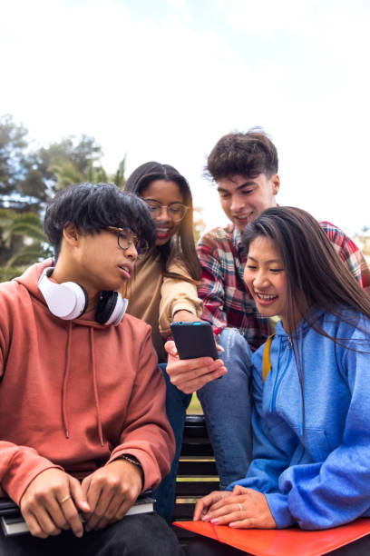 Multiracial happy young college student friends look at mobile phone. Teenagers using smartphone outdoor. Vertical. Group of multiracial happy young college student friends look at mobile phone. Teenagers using smartphone together outdoor. Vertical image. Youth lifestyle and social media addiction concept. education student mobile phone university stock pictures, royalty-free photos & images