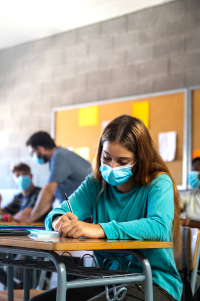istock High school female student wearing face mask in class.doing homework. Teacher helping student in background. Vertical. 1423903599