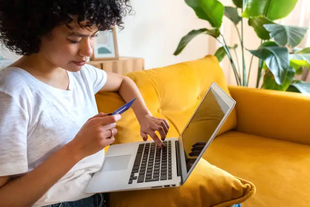 Close up of african american woman sitting on the couch using credit card and laptop to shop online from home. Copy space. E-commerce concept.