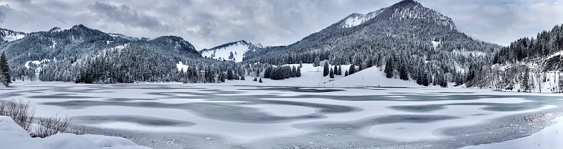 Panoramic view of the winter lake Spitzingsee. A wide photo of the snow-covered lake Spitzingsee. Alpine mountains beyond the lake in winter. A frozen lake in Bavaria surrounded by the Alps.