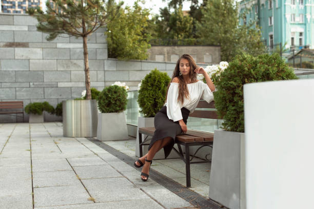 Young woman with curly hair sits on a white and wooden bench in a park, enjoying the peaceful surroundings and looking away from the camera