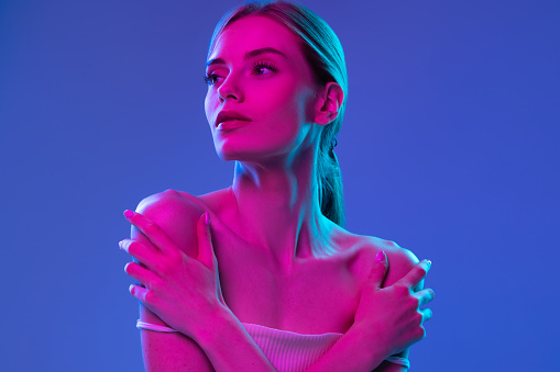 Inspiration. Young beautiful fashion model with well-kept skin and naked shoulders isolated over dark background in pink neon light. Vivid style, beauty, art, future concept.