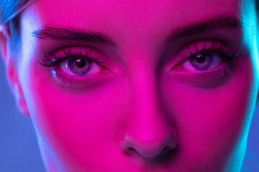 Blue eyes. Closeup eyes of beautiful young girl looking at camera in pink neon light. Concept of cosmetics, makeup, natural and eco treatment, skin care. Shiny and healthy look, fashion, vision. Details