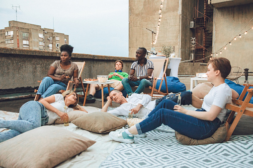 A multiracial group of tenants enjoys their time while watching a movie on the rooftop at dusk. In this photo, they are sitting or lying down.