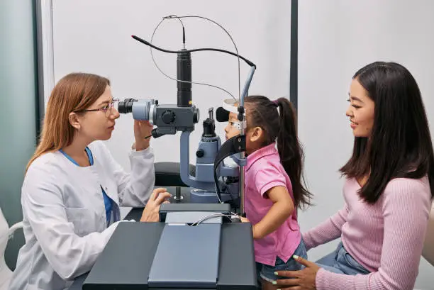 Japanese mother with her daughter while visit to optometrist. Little female child getting vision testing and eye exam in ophthalmology clinic, side view