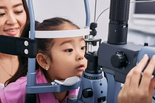 Checking little Korean girl's eyesight with binocular slit lamp in ophthalmology clinic, close-up. Vision correction in children