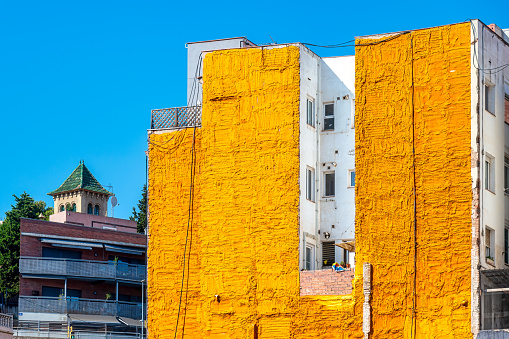 Barcelona, Spain - August 25, 2022: View of a white-story building smeared with a thick layer of yellow paint at the front.  Another building is beside the story building with trees in the background.