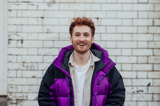 A young redhead man looking at the camera, getting his portrait taken against a white bricked wall. He is wearing a purple winter jacket.