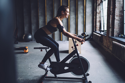 Side view of young sporty female in activewear exercising on stationary bike in loft styled gym and listening to music through earphones