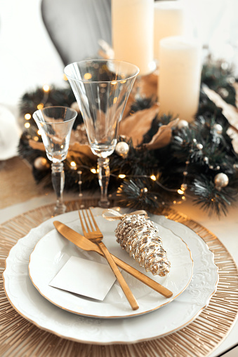 Closeup table xmas decor with fir wreath two plates, golden cutlery, crystal glasses, with blank guest card and a Christmas toy.