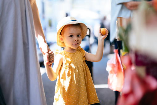 Close up shot of a cute little girl in a yellow dress and a hat, holding anonymous woman's hand while they are walking through the green market. The girl is picking fruits.