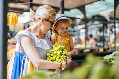 Portrait of a beautiful woman wearing glasses is holding her cute little daughter in her arms while they are picking lettuce at the green market. They are smiling and looking down.