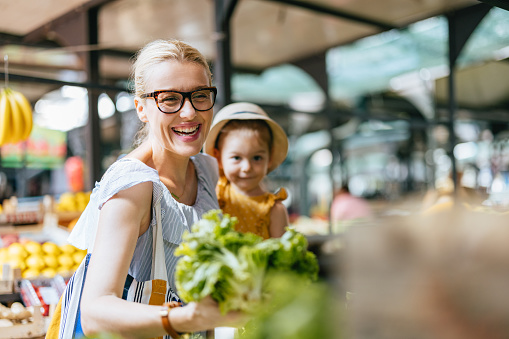 Portrait of a beautiful woman wearing glasses is holding her cute little daughter in her arms while they are picking lettuce at the green market. They are smiling and looking down.
